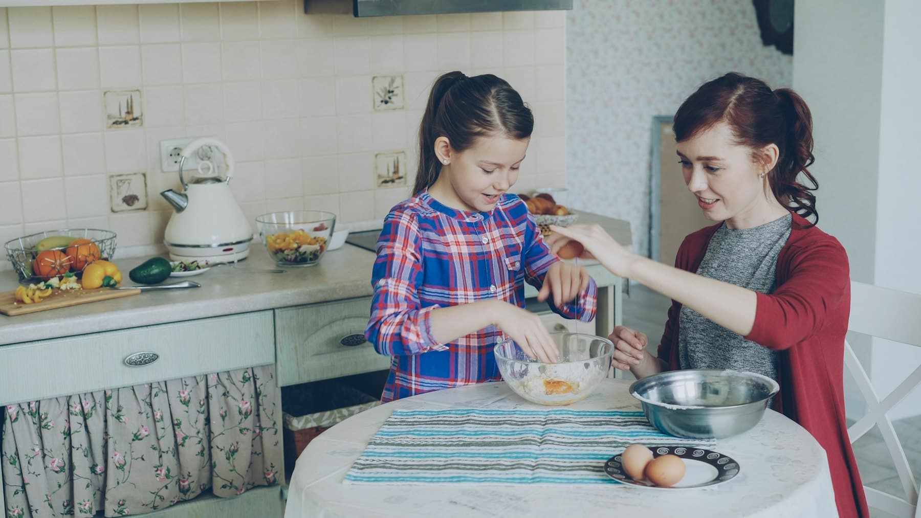 A young girl mixing ingredients in a home kitchen with help from a family member
